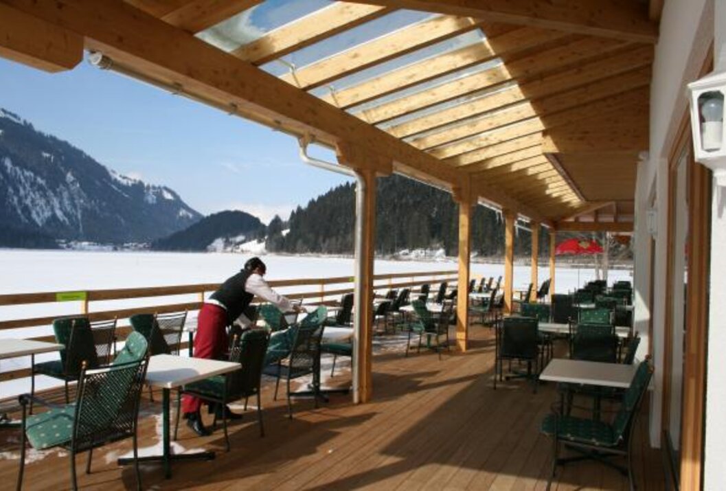 Kellnerin deckt Tische auf einer sonnigen Terrasse mit Blick auf einen verschneiten See und Berge