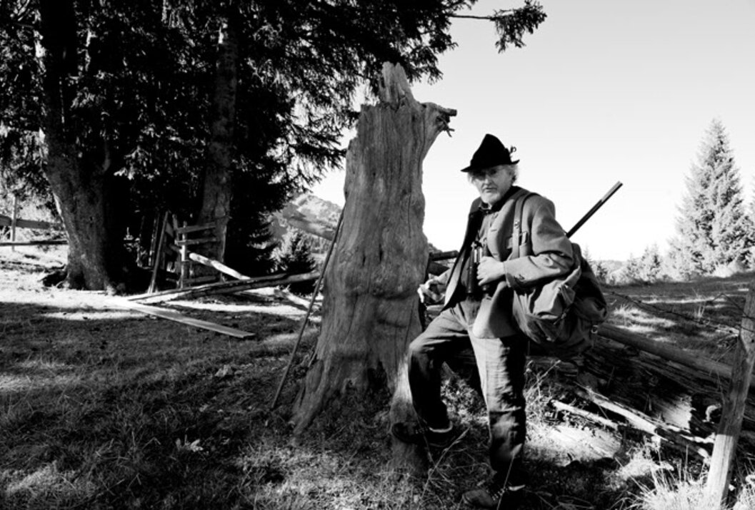 Schwarzweißfoto eines Mannes mit Hut und Gewehr neben einem Baumstumpf