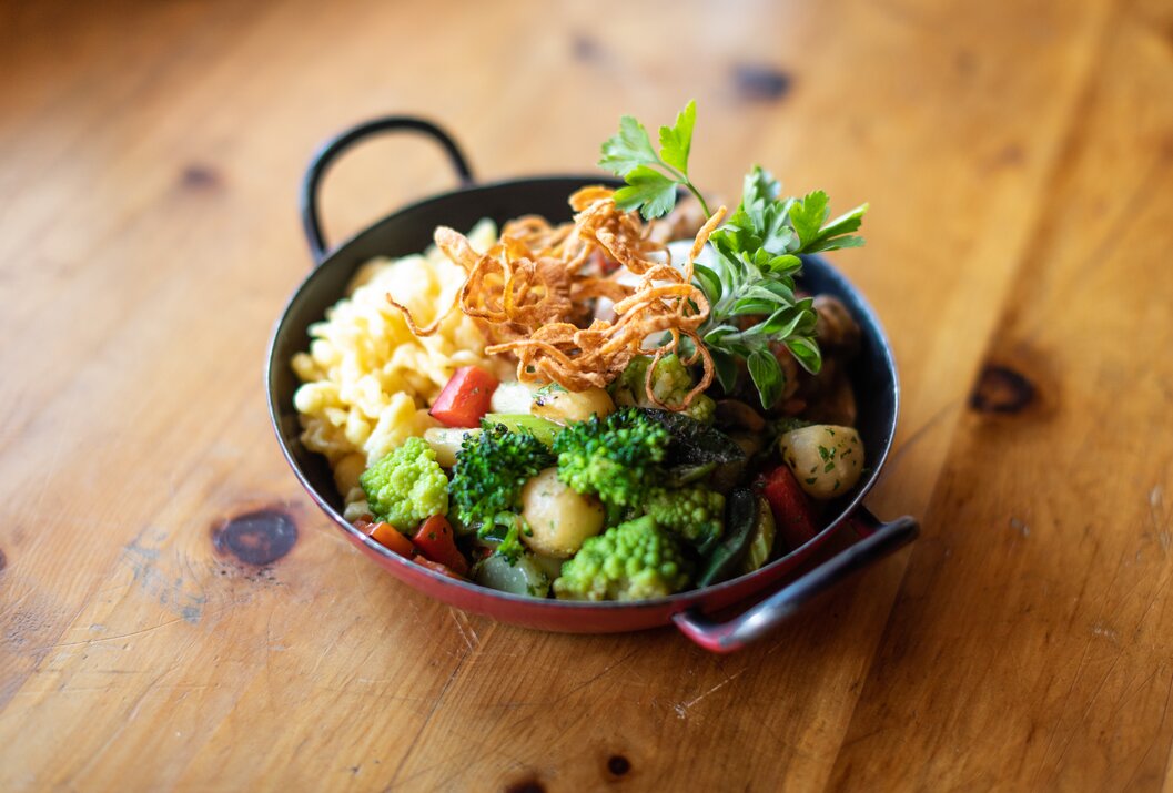 Käsespätzle mit Gemüse und Röstzwiebeln in einer roten Pfanne auf einem Holztisch