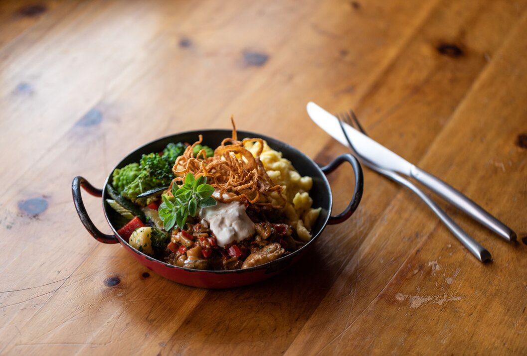 Schüssel mit Gemüse, Spätzle, Fleisch und Soße auf Holztisch, daneben Messer und Gabel