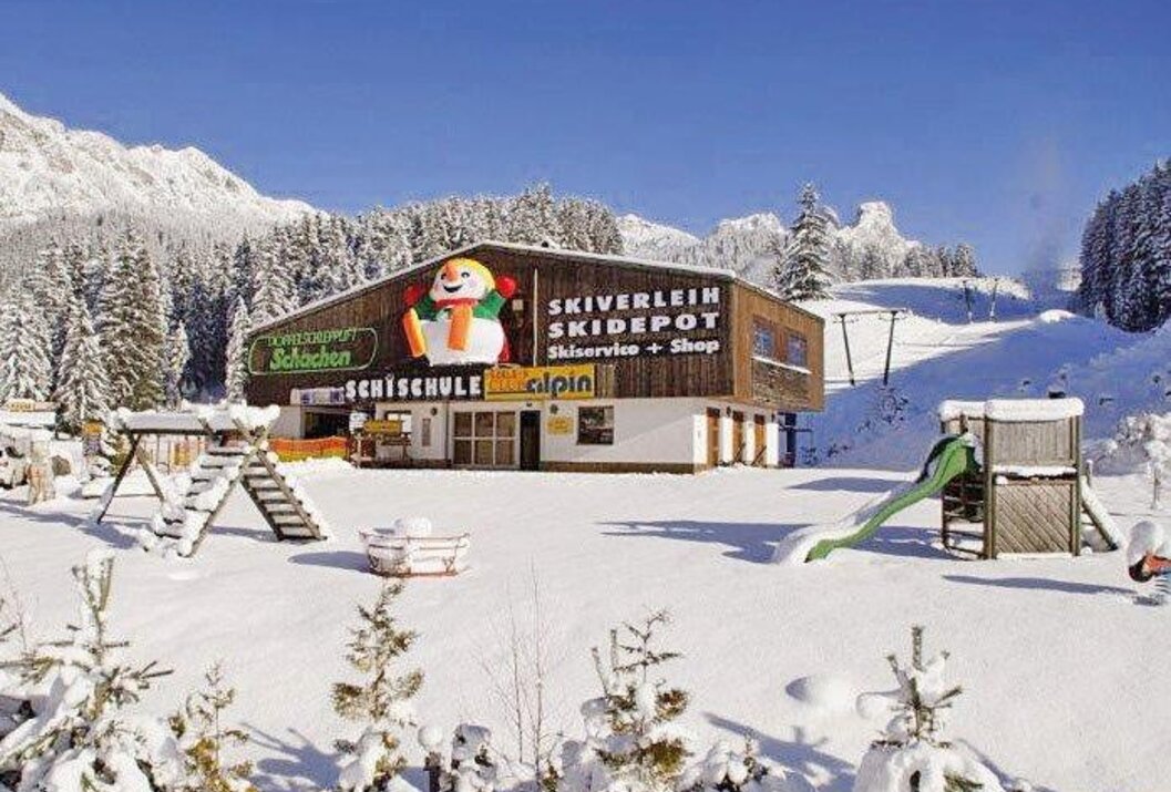 Skischule im Schnee mit Schneemann-Logo, Rutsche und Bergen im Hintergrund