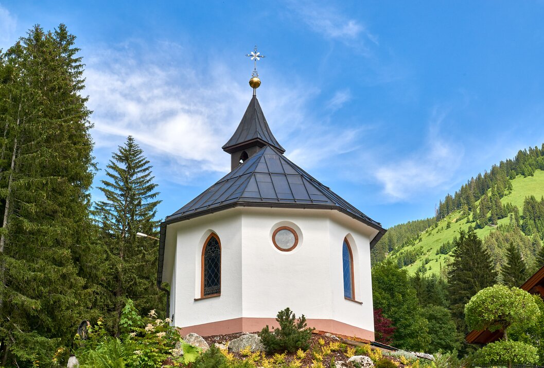 Kleine weiße Kapelle mit schwarzem Dach vor grünem Hügel und blauem Himmel