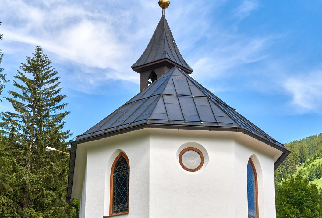 Kleine weiße Kapelle mit dunklem Dach vor blauem Himmel und grüner Landschaft