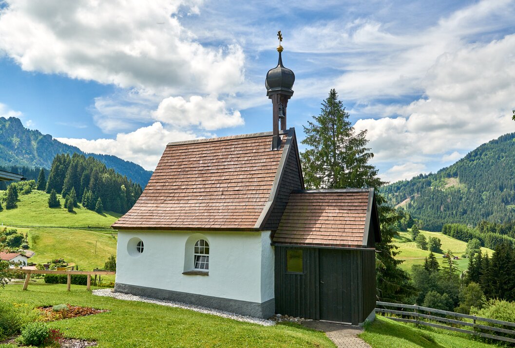 Kleine weiße Kapelle mit braunem Dach vor grüner Landschaft und blauem Himmel mit Wolken
