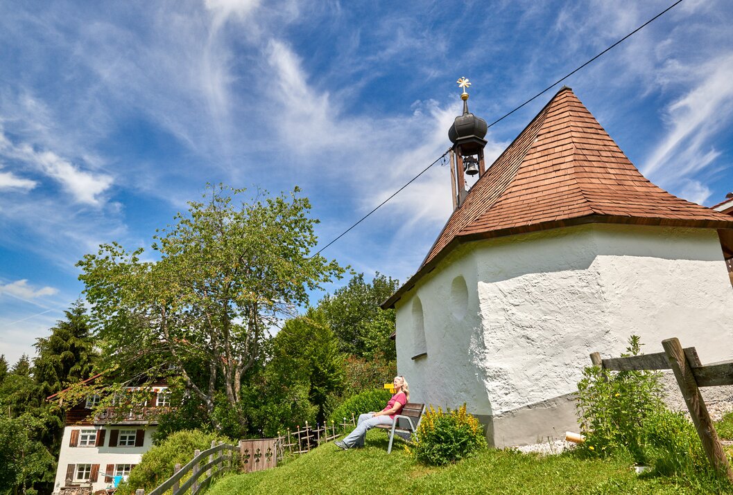 Frau sitzt auf Bank vor kleiner Kapelle mit rotem Dach und Kirchturm unter blauem Himmel