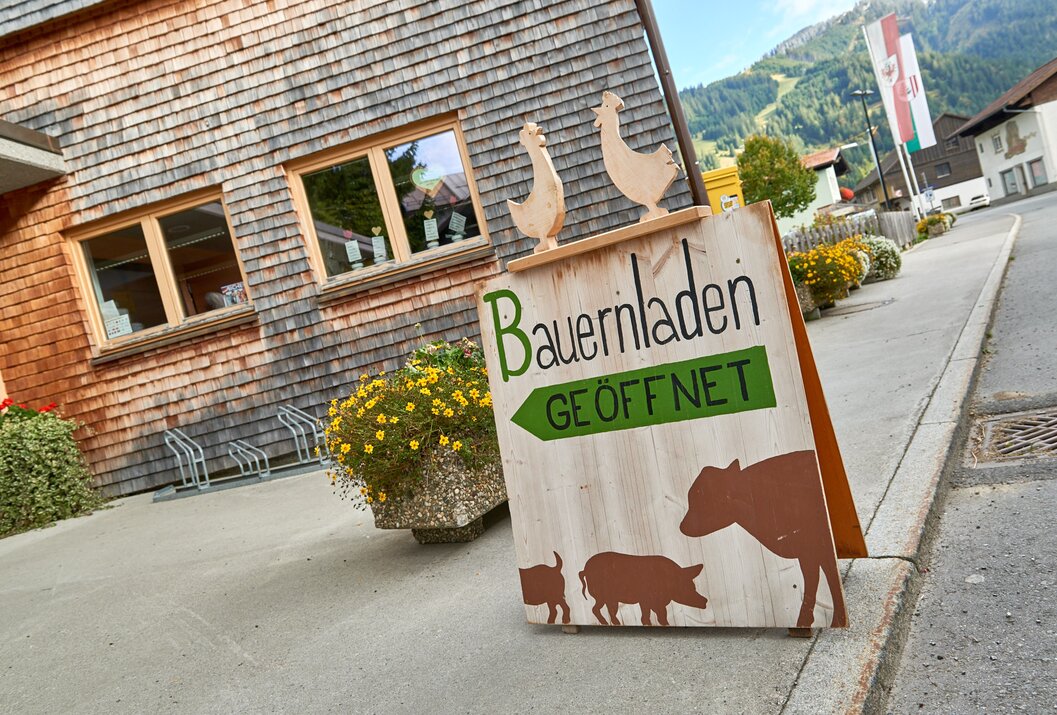 Schild "Bauernladen GEÖFFNET" mit Kuh, Schwein und Ferkel vor Holzhaus mit Fenster und Fahrrädern