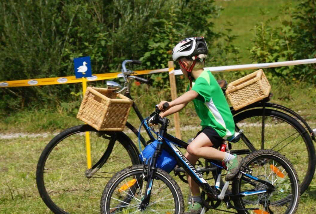 Boy with helmet riding a bicycle with a basket, next to another bicycle, green in the background