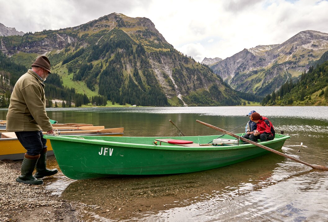 Grünes Boot am Ufer eines Sees, im Hintergrund Berge und zwei Personen im Boot