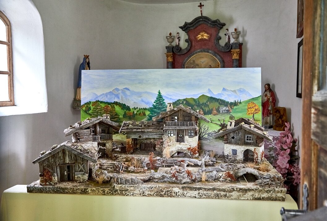 Krippe mit Häusern vor gemalter Berglandschaft, darüber religiöse Figuren und ein Kreuz