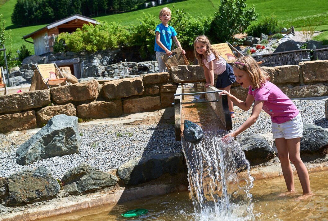 Drei Kinder spielen an einem Wasserlauf mit Steinen und Holzrinnen im Freien