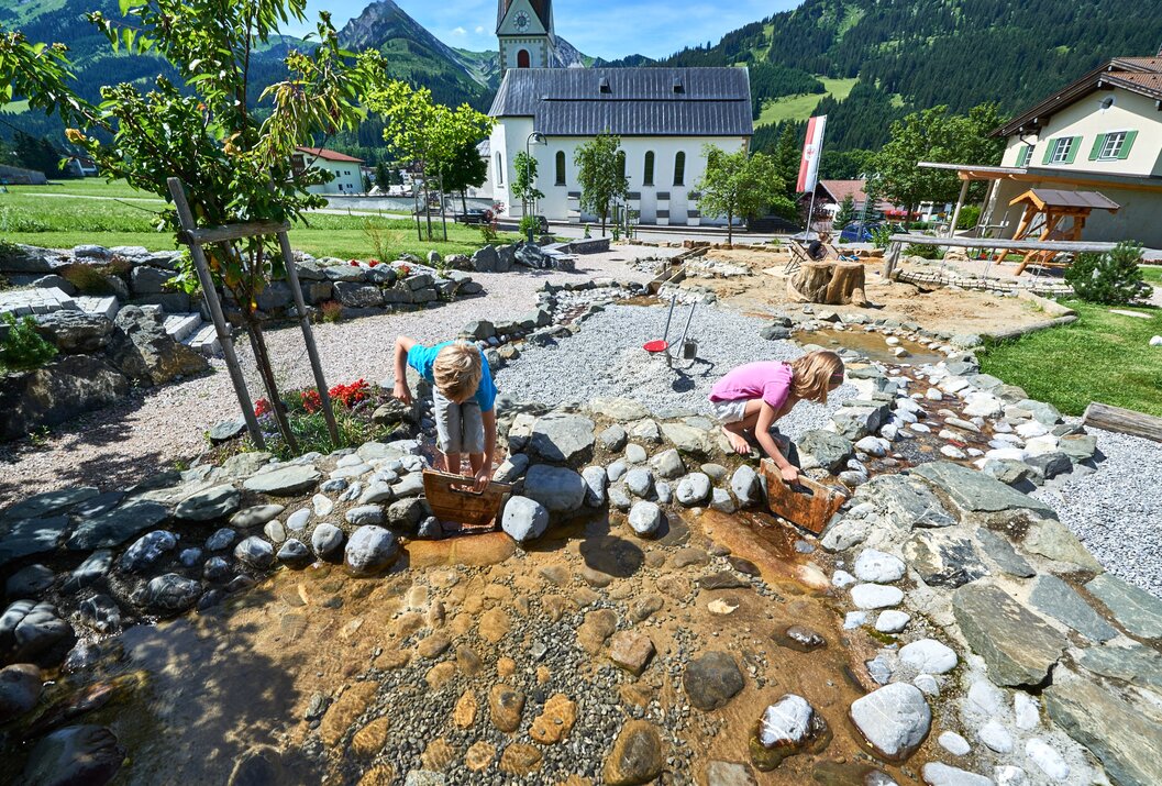 Zwei Kinder spielen an einem Bachlauf mit Steinen und Holzeimern vor einer Kirche in den Bergen
