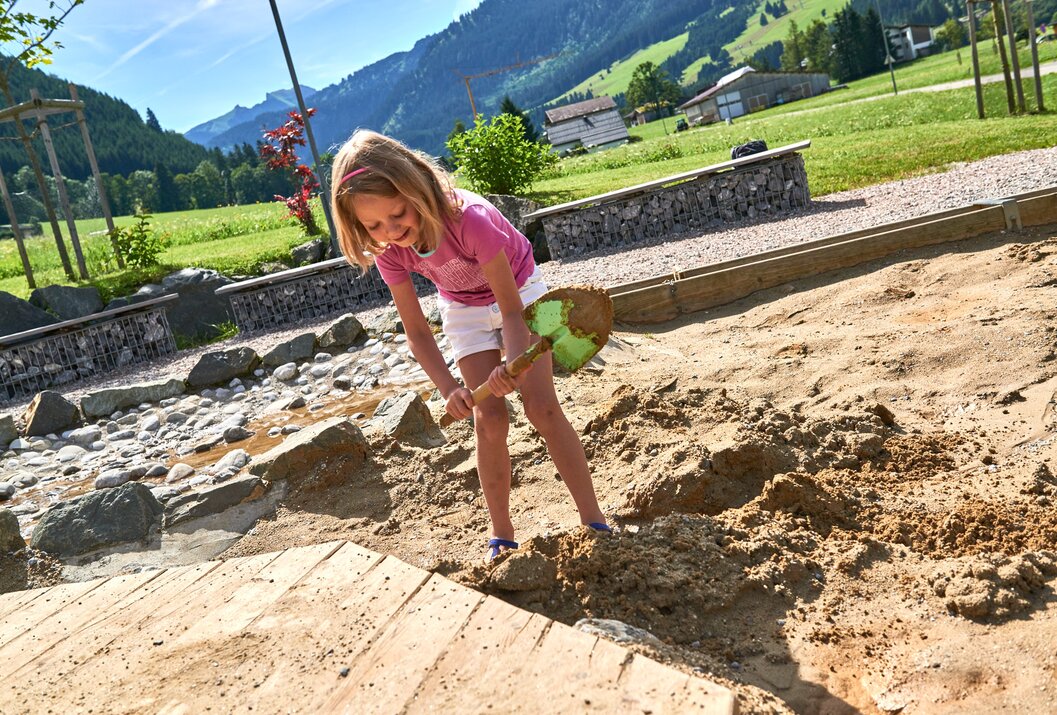 Mädchen schaufelt Sand auf einem Spielplatz vor Bergkulisse