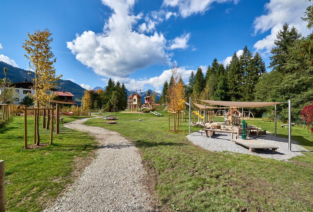 Spielplatz mit Holzhäusern, Sandkasten und Bäumen vor Bergkulisse unter blauem Himmel