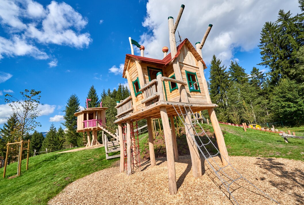 Hölzerner Spielplatz mit Baumhaus und Kletternetz auf einem Hügel unter blauem Himmel
