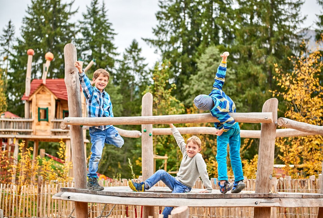 Drei Jungen spielen auf einem hölzernen Klettergerüst im Freien vor Bäumen
