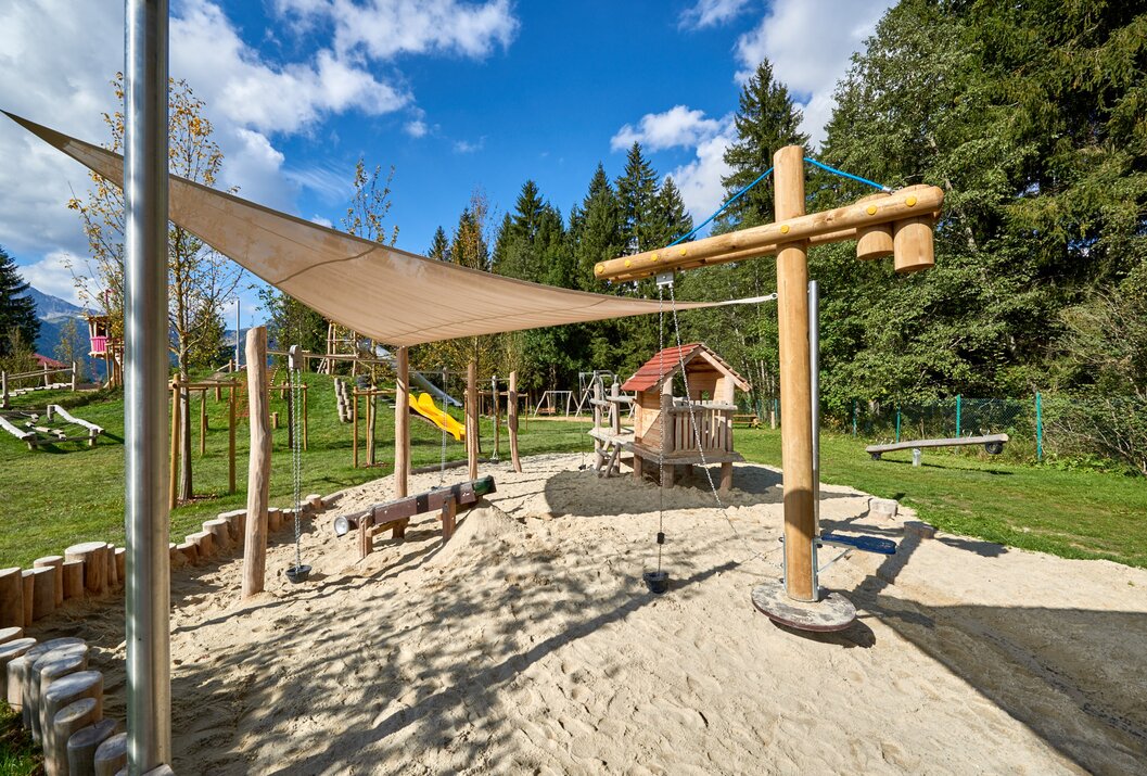 Spielplatz mit Sand, Holzelementen, Sonnensegel und Spielhaus vor Bäumen und blauem Himmel
