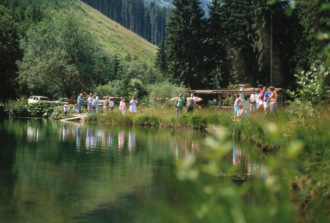 Menschengruppe am Ufer eines Sees vor bewaldeten Bergen