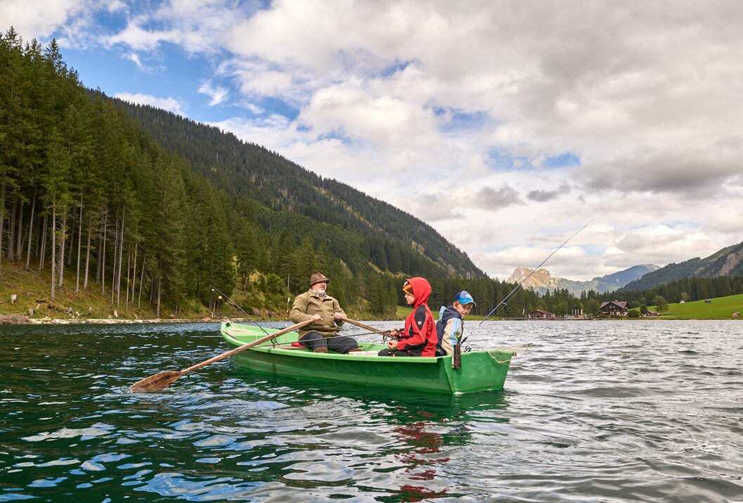 Mann und zwei Kinder angeln in einem grünen Ruderboot auf einem See vor bewaldeten Bergen