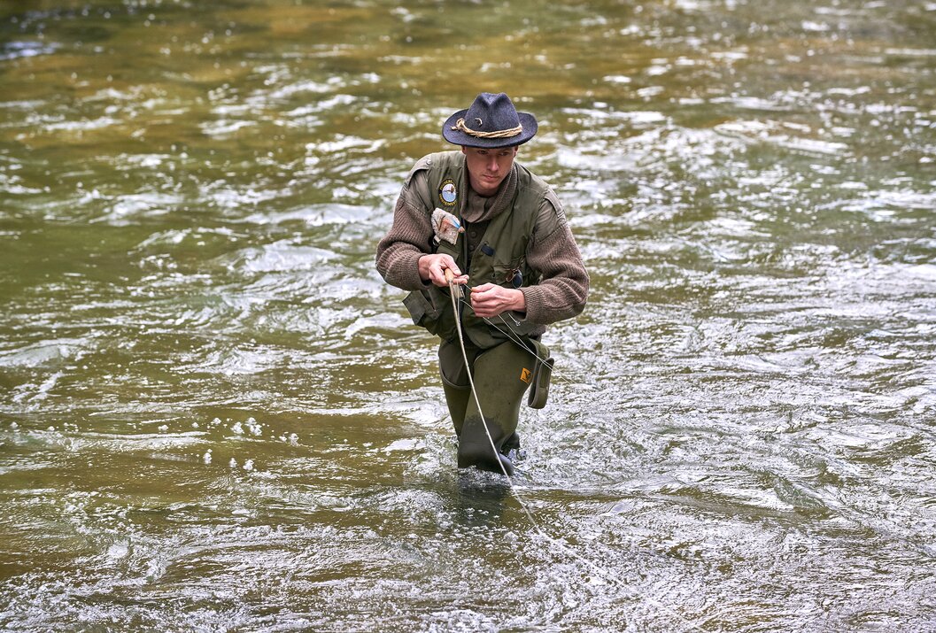 Mann mit Hut und Wathose steht im Fluss und angelt