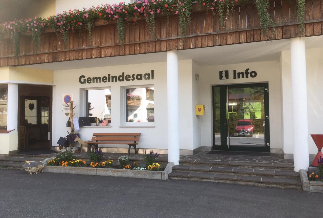 Gebäude mit Aufschrift Gemeindesaal und Info, Blumenkästen und Bank vor dem Eingang