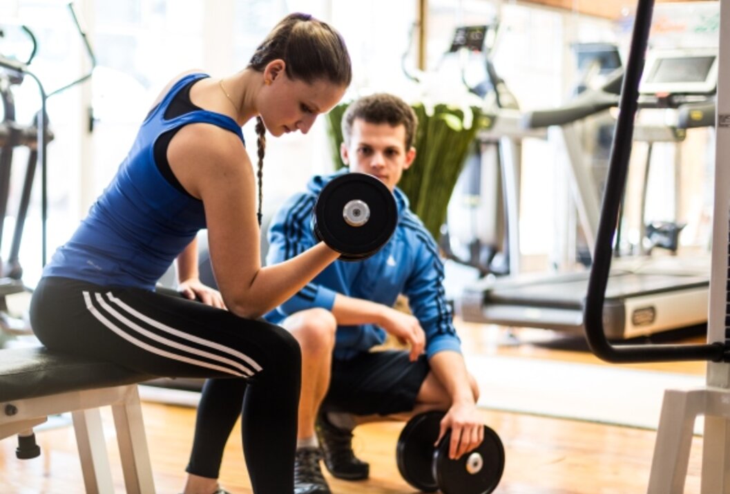 Frau trainiert Bizeps mit Hantel, Mann beobachtet im Fitnessstudio
