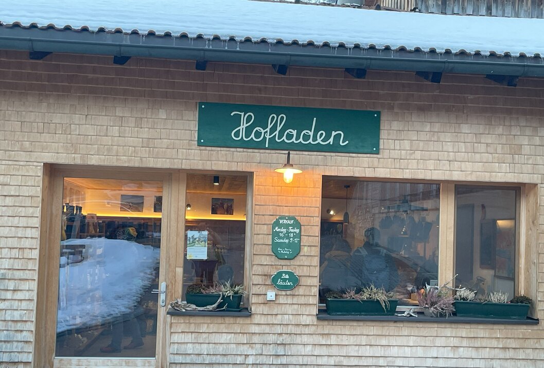 Holzfassade eines Hofladens mit grünem Schild und Öffnungszeiten