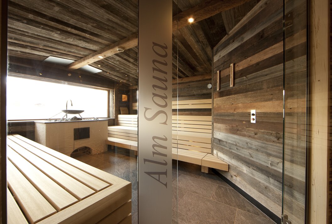 Holzsauna mit Bänken, Ofen und Fenster, abgetrennt durch Glaswand mit Aufschrift Alm Sauna