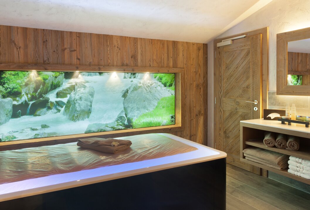 Innenraum mit Liege, Holzwand und Fensterblick auf einen Wasserfall