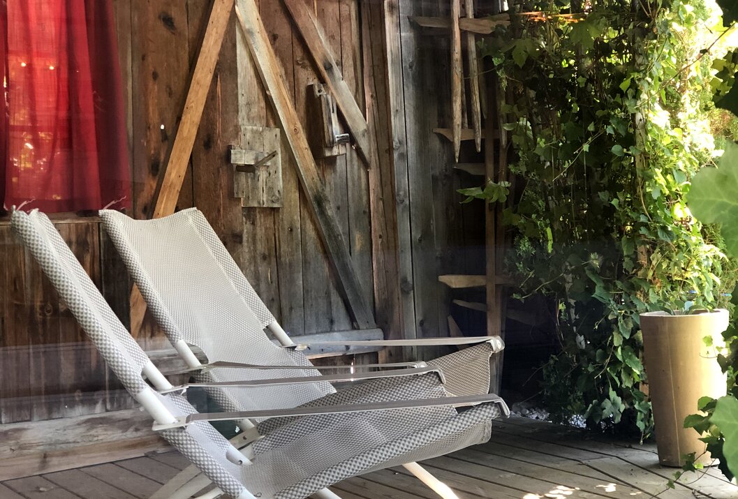 Zwei Liegestühle auf Holzterrasse vor Holztür, Rankpflanzen und Leiter im Hintergrund