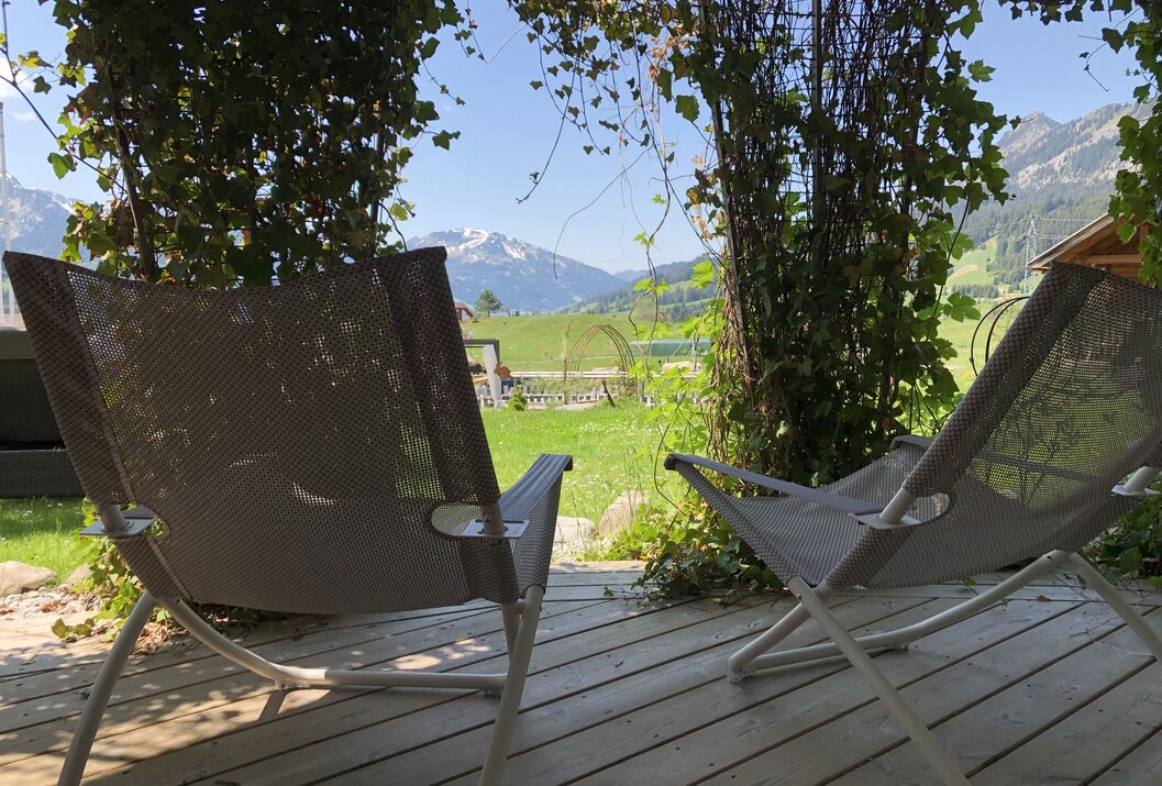 Zwei Liegestühle auf Holzterrasse mit Blick auf grüne Wiese und schneebedeckten Berg
