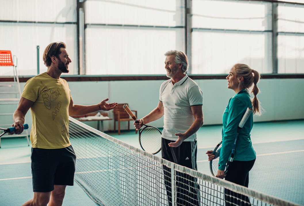 Drei Personen mit Tennisschlägern stehen am Netz und unterhalten sich
