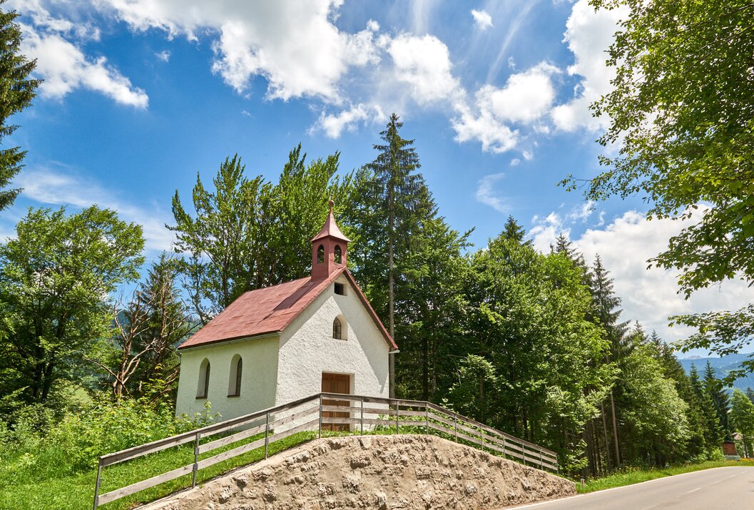 Kleine weiße Kapelle mit rotem Dach vor grünem Wald und blauem Himmel mit Wolken