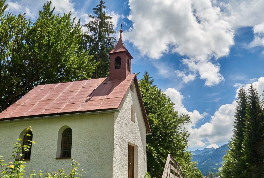 Kleine weiße Kapelle mit rotem Dach vor grünen Bäumen und blauem Himmel mit Wolken