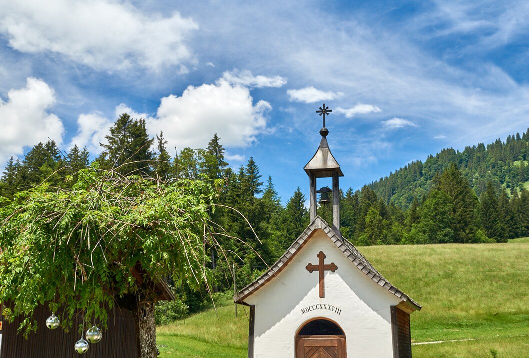 Kleine Kapelle mit Holztür vor grüner Wiese und bewaldetem Hügel unter blauem Himmel mit Wolken
