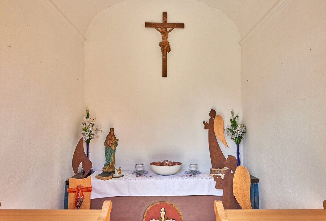 Kleine Kapelle mit Kruzifix, Heiligenfigur, Engeln und Blumen auf einem Altar mit weißer Decke