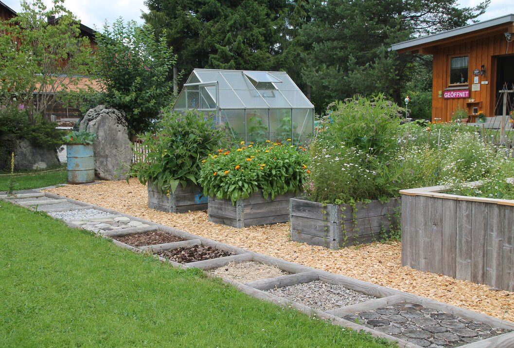 Garten mit Hochbeeten, Gewächshaus und Weg mit verschiedenen Materialien