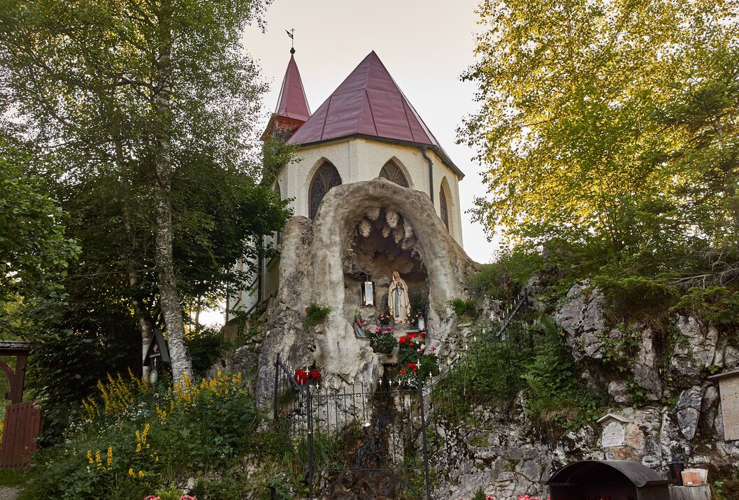 Kleine Kirche mit rotem Dach und Marienstatue in Felsengrotte zwischen Bäumen