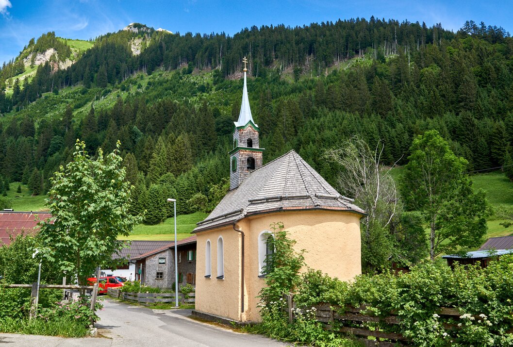 Kleine Kirche mit grünem Turm vor bewaldetem Berghang