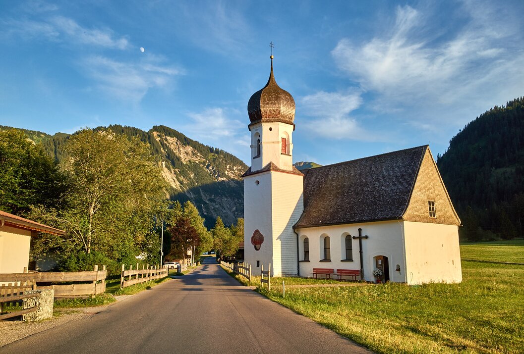 Weiße Kirche mit Zwiebelturm vor Bergkulisse an Straße mit Holzzaun
