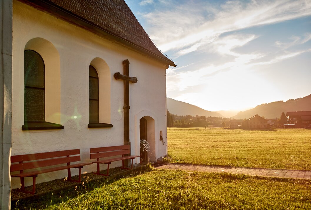 Kleine weiße Kirche mit Holzkreuz vor Wiese und Bergen im Sonnenlicht
