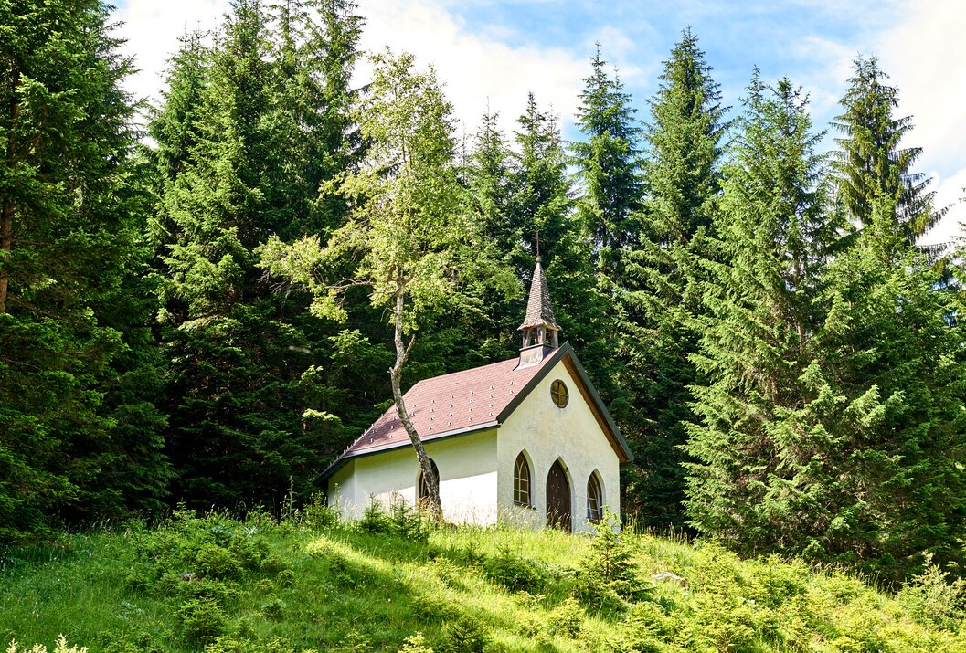 Kleine Kapelle mit rotem Dach am Waldrand auf grüner Wiese
