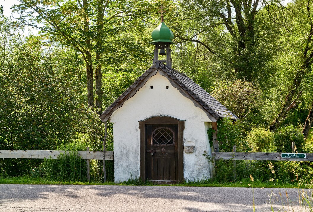 Kleine weiße Kapelle mit grünem Turm und Holztür vor Bäumen und Zaun