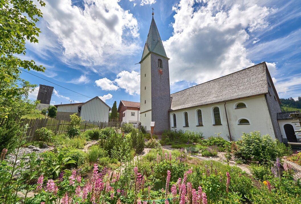 Kirche mit hohem Turm, umgeben von Blumen und Grün unter blauem Himmel mit Wolken