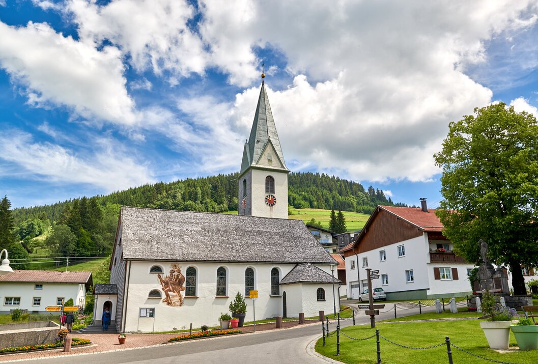 Kirche mit hohem Turm in ländlicher Umgebung vor bewaldetem Hügel und blauem Himmel mit Wolken