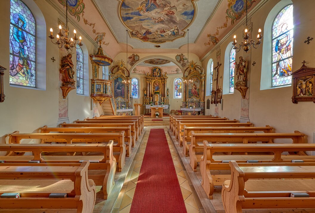 Innenansicht einer Kirche mit Holzbänken, rotem Teppich und Deckenmalerei