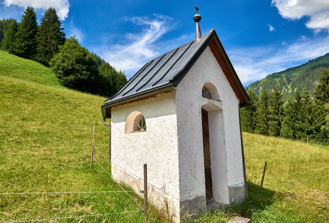 Kleine weiße Kapelle auf grüner Wiese vor bewaldetem Hügel unter blauem Himmel