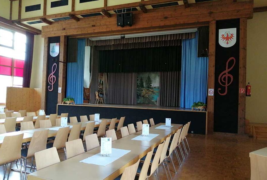 Bühne mit Vorhang und Waldkulisse, davor lange Tische mit Stühlen in einem Saal