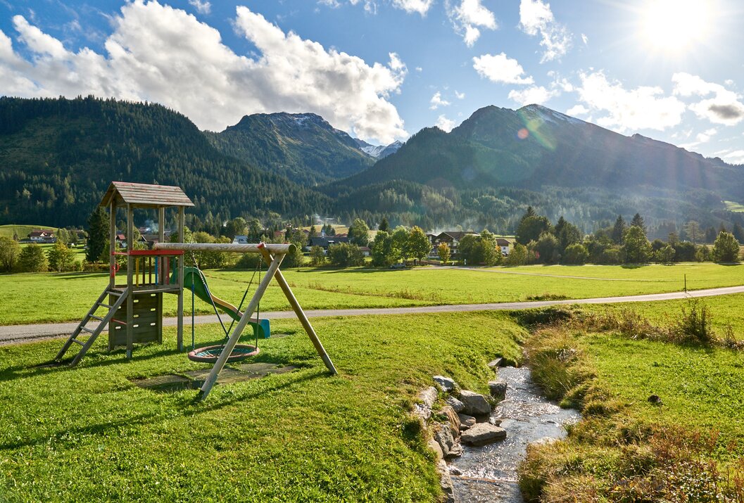 Spielplatz vor Bergkulisse mit Wiese und Bachlauf im Vordergrund