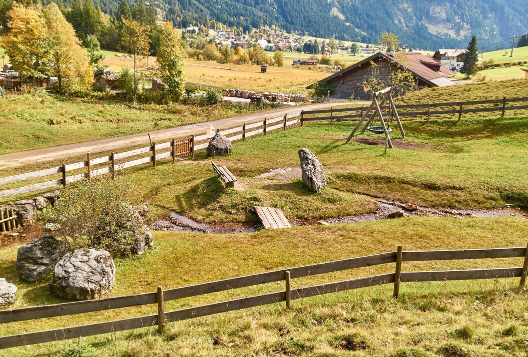 Ländliche Szene mit Holzzaun, Bachlauf, Spielplatz und Häusern vor bergiger Landschaft