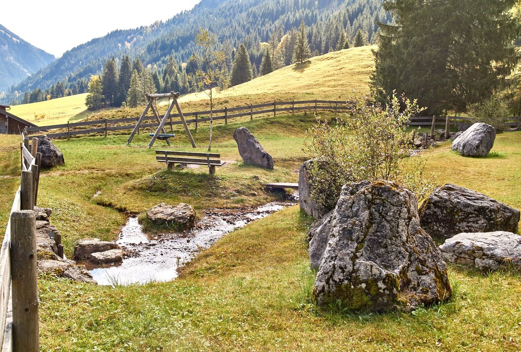 Idyllische Landschaft mit Bach, Schaukel, Bank und Felsen vor bewaldetem Berghang